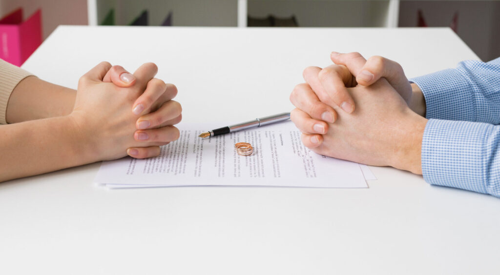 Two people sitting opposite of one another with their hands clasped separately over a paper with a pair of rings.